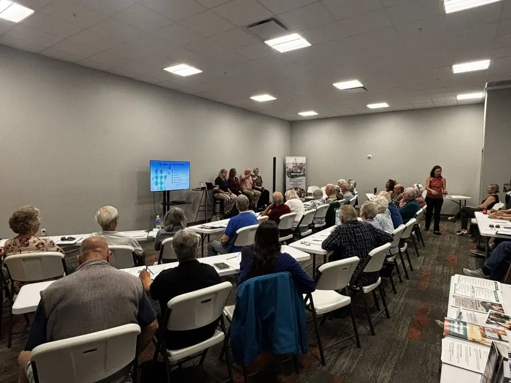 tables are filled with older adults at a lifelong learning for seniors program with 4 speakers at the front