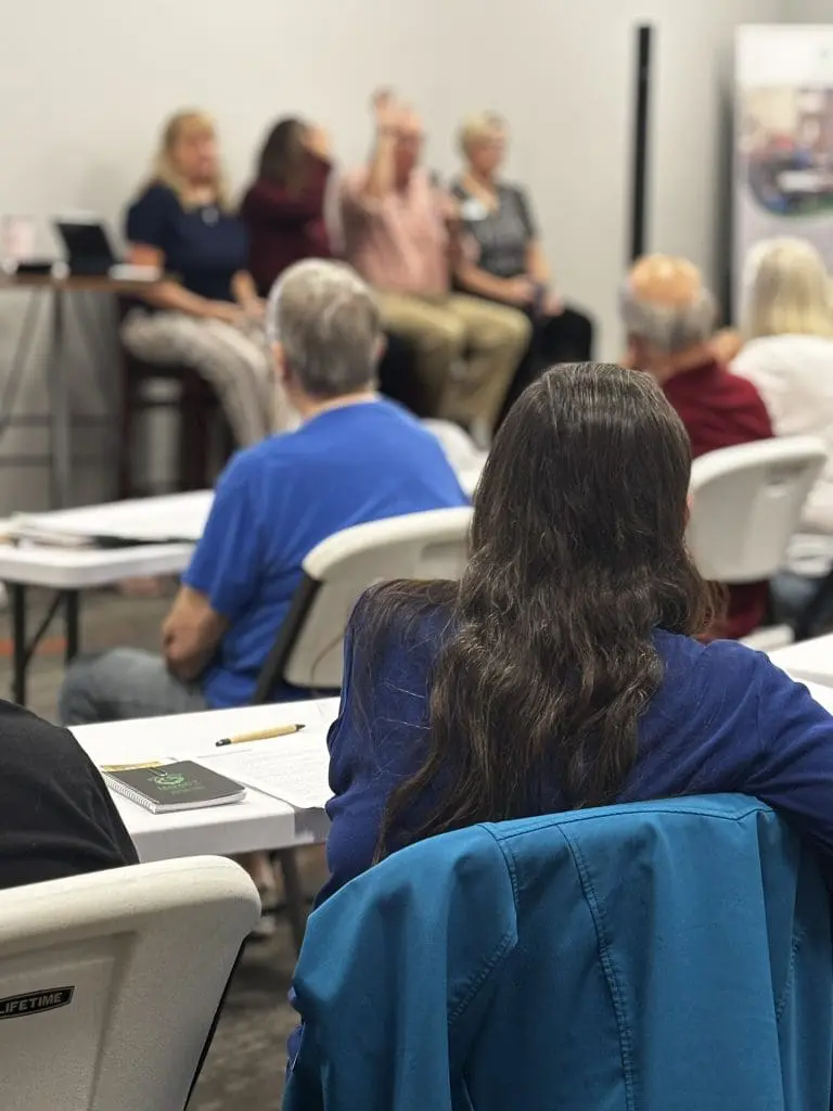 A woman with dark hair and a blue shirt is seated looking away from the camera at 4 panelist at an adult learning seminar